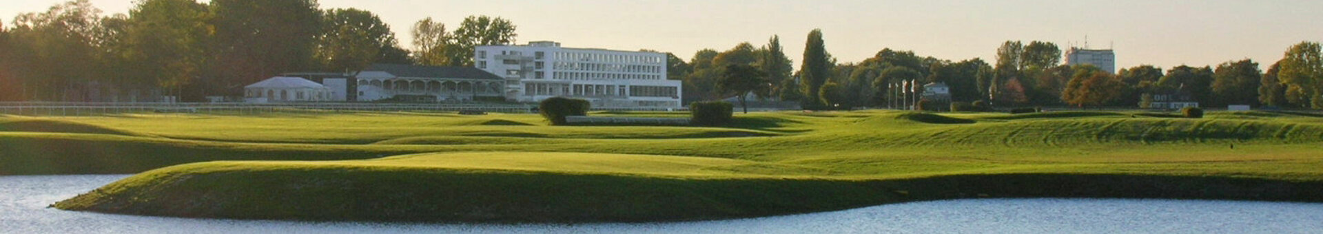 Grüne Wiese mit Wassergraben, dahinter das ATLANTIC Hotel Galopprennbahn in Bremen unter blauem Himmel.