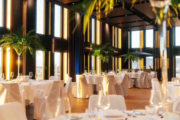 Elegantes Hotelrestaurant mit hohen Fenstern, weißen Tischdecken und Palmen, bietet eine luxuriöse und helle Atmosphäre.