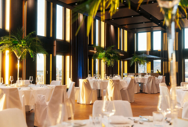 Elegantes Hotelrestaurant mit hohen Fenstern, weißen Tischdecken und Palmen, bietet eine luxuriöse und helle Atmosphäre.