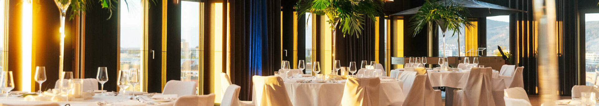 ATLANTIC Hotel Heidelberg Ballsaal Firmenfeier Elegant hotel restaurant with high windows, white tablecloths and palm trees, offering a luxurious and bright atmosphere.