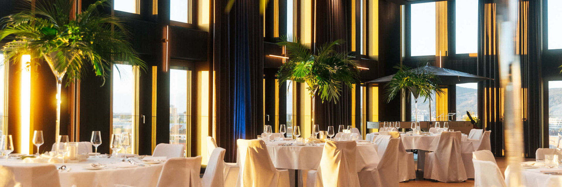 Elegant hotel restaurant with high windows, white tablecloths and palm trees, offering a luxurious and bright atmosphere.