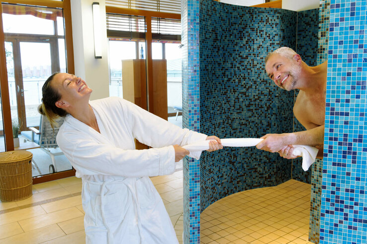 A laughing couple play happily with a towel in a wellness area with blue mosaic tiles.