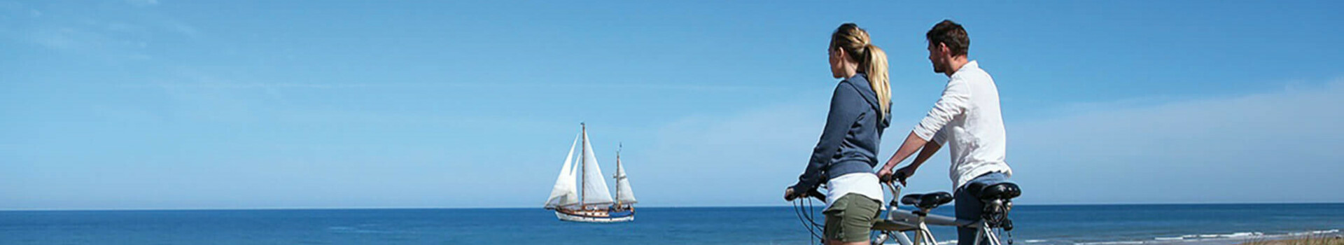 Aktion Sail & Bike Couple with bicycles looking from a dune to the sea, sailing ship on the horizon under a blue sky.