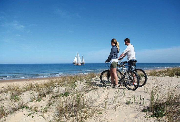 Paar mit Fahrrädern blickt von einer Düne auf das Meer, segelndes Schiff am Horizont unter blauem Himmel.