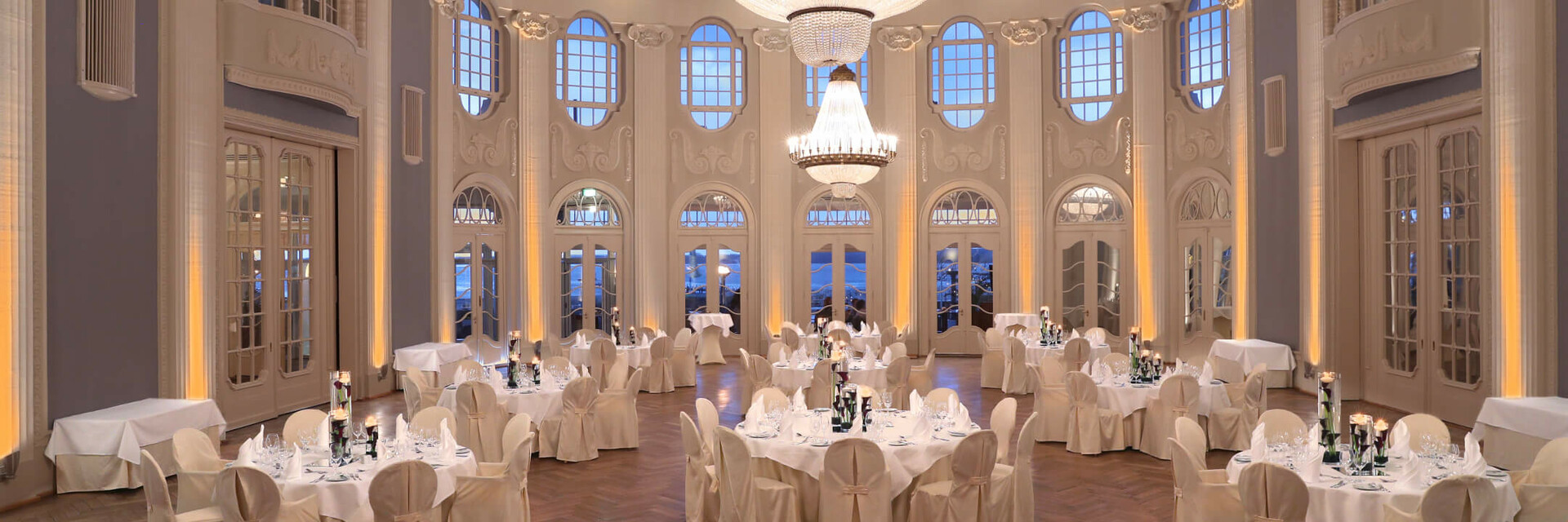 Elegant ballroom in the ATLANTIC Grand Hotel Travemünde with chandelier, round tables and white chairs.