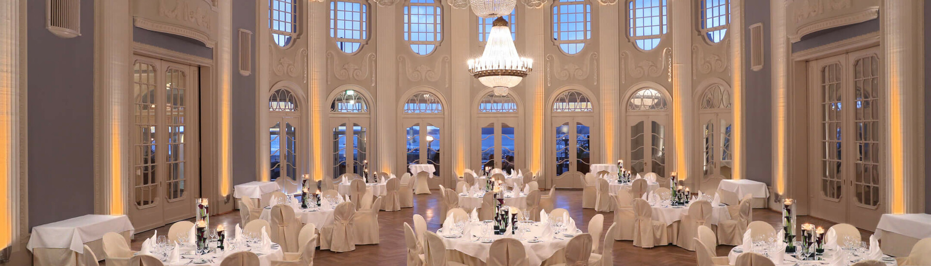 Elegant ballroom in the ATLANTIC Grand Hotel Travemünde with chandelier, round tables and white chairs.