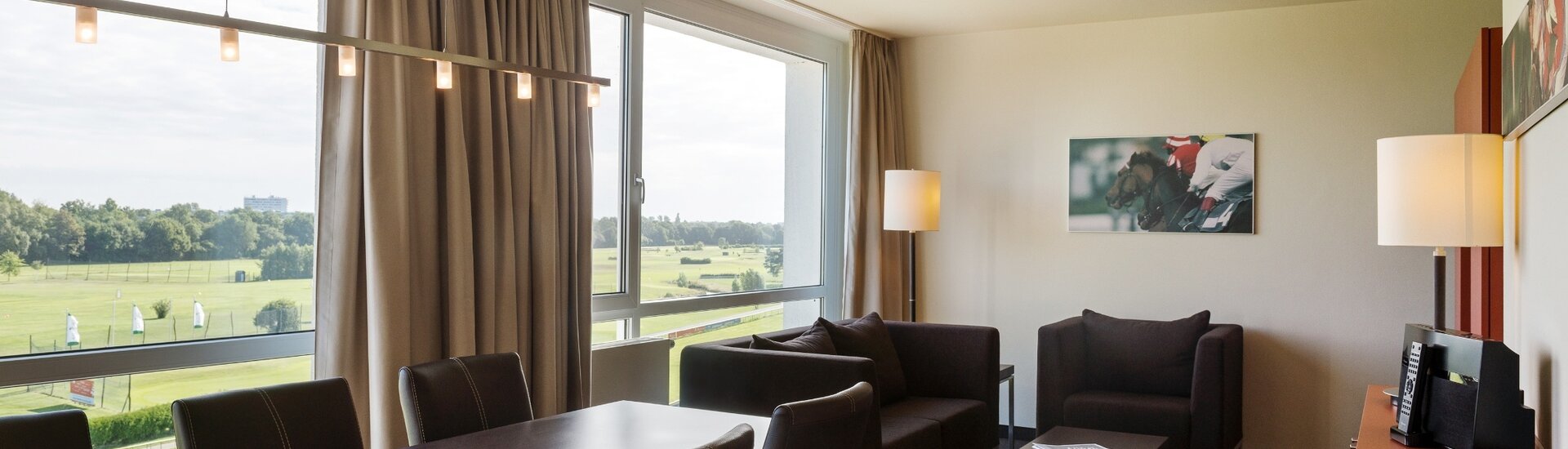 Living area of the Studio in the ATLANTIC Hotel Galopprennbahn Bremen  Bright hotel room with large window, view of the racecourse, dining table, chairs, sofa and modern lighting.