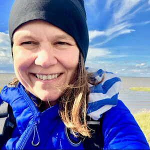 Person in blauer Jacke lächelt am Meer unter blauem Himmel.
