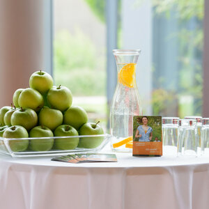 Ein Tisch mit grünen Äpfeln, Wasserkaraffe mit Orangen, Gläsern und Broschüren in einem hellen Raum.