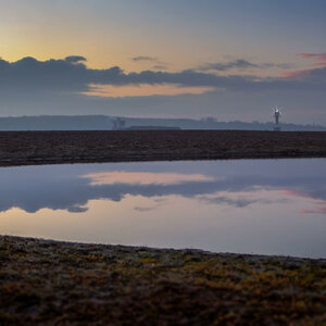Sonnenaufgang am Strand mit ruhigem Wasser und Leuchtturm im Hintergrund.