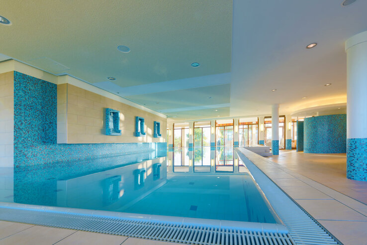 Bright indoor pool with blue tiled walls, large windows and comfortable seating areas.