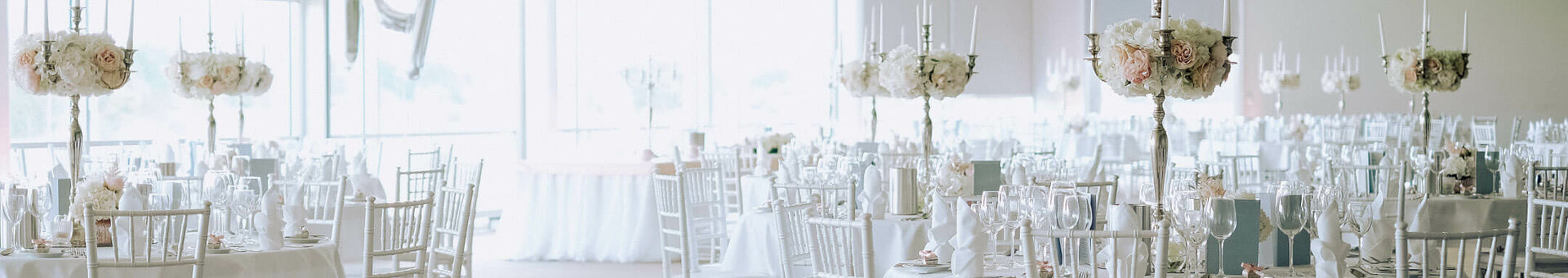 Elegant, bright banquet hall with white-clothed tables, flower arrangements and Chiavari chairs.