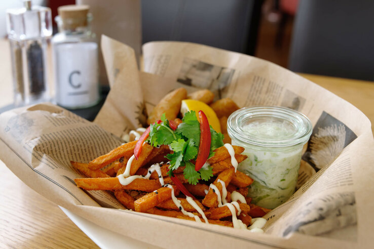 Süßkartoffelpommes mit Mayonnaise, Koriander und Paprika, serviert mit Dip im Glas, auf Zeitungspapier angerichtet.