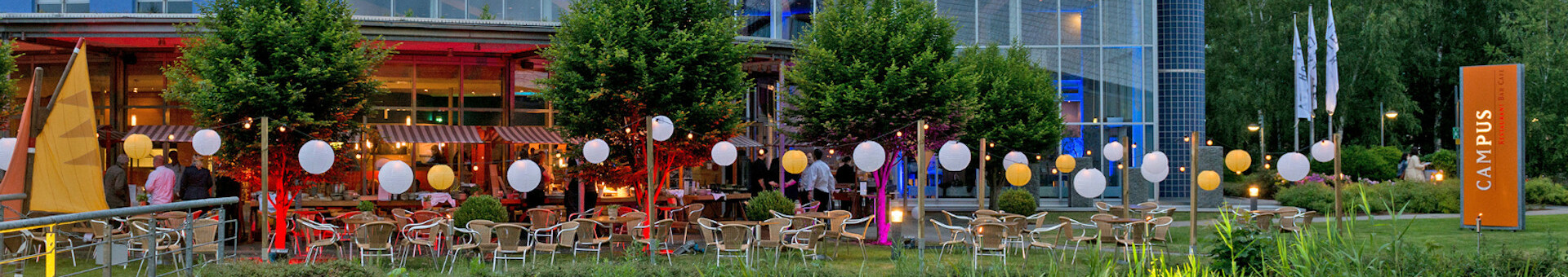 Illuminated terrace of the ATLANTIC Hotel Universum Bremen with tables, chairs and lanterns at dusk.