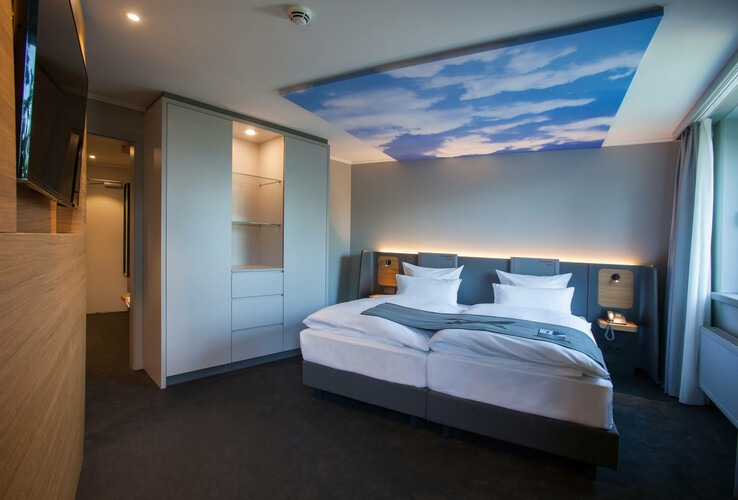 Modern hotel room with double bed, stylish furniture, large TV and ceiling with sky motif at the ATLANTIC Hotel Airport Bremen.