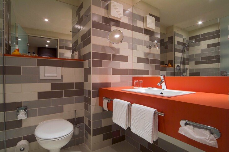 Modernes Badezimmer mit orangefarbenem Waschtisch, Spiegel, Handtüchern und Dusche im ATLANTIC Hotel Galopprennbahn Bremen.