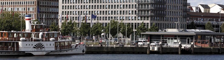 Direkt an der Kieler Förde gelegen – Außenansicht des ATLANTIC Hotel Kiel bei Tag Blick auf das ATLANTIC Hotel Kiel von der Förde aus, mit einem Schiff im Vordergrund und blauem Himmel.
