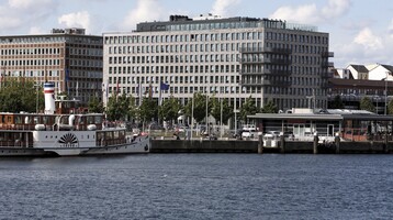 Blick auf das ATLANTIC Hotel Kiel von der Förde aus, mit einem Schiff im Vordergrund und blauem Himmel.