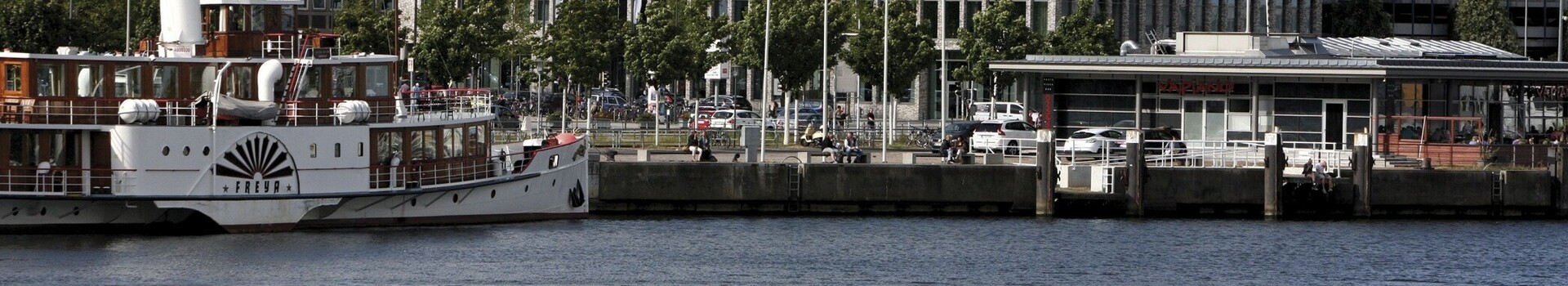 Direkt an der Kieler Förde gelegen – Außenansicht des ATLANTIC Hotel Kiel bei Tag Blick auf das ATLANTIC Hotel Kiel von der Förde aus, mit einem Schiff im Vordergrund und blauem Himmel.