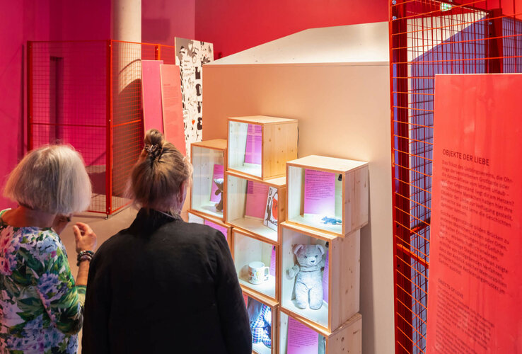 Zwei Personen betrachten beleuchtete Ausstellungsboxen mit persönlichen Objekten in einem farbenfrohen Museumsraum.