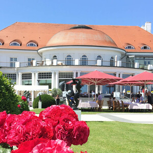 Historische Hotelansicht mit rotem Dach, Terrasse, roten Sonnenschirmen und blühenden Rosen im Vordergrund.