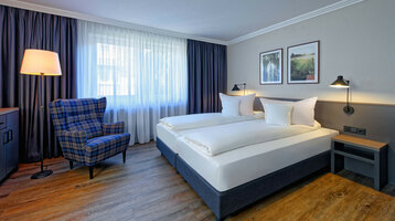 Rooms at the ATLANTIC Hotel Landgut Horn Bremen Bright hotel room with double bed, checkered armchair, floor lamp, wooden floor and pictures on the wall.