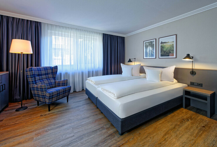 Helles Hotelzimmer mit Doppelbett, kariertem Sessel, Stehlampe, Holzboden und Bildern an der Wand.