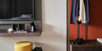 Hotelzimmer mit TV, Regal, Lampe, gelbem Hocker, Aktentasche, Kleiderstange mit Jacke und Pullover, Schuhe auf Bank.