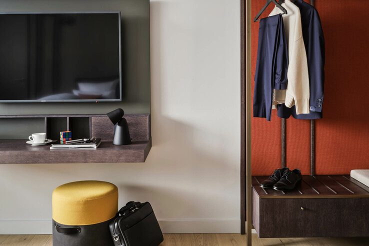 Hotel room with TV, shelf, lamp, yellow stool, briefcase, clothes rail with jacket and sweater, shoes on bench.