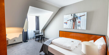 Helles Hotelzimmer mit Einzelbett, modernem Sessel, kleinem Tisch und Bild von einem Schiffsmast an der Wand.