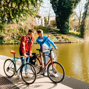 Two people with bicycles are standing on the shore of a lake, surrounded by green nature and trees.