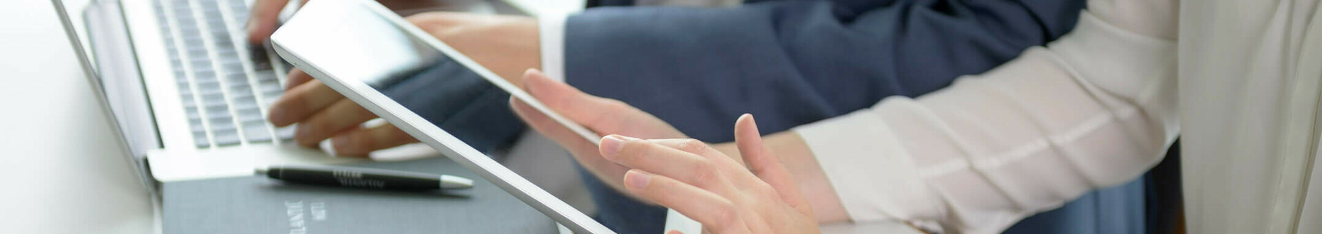 Smarte Media-Nutzung in den ATLANTIC Hotels Zwei Personen arbeiten an Laptop und Tablet in moderner Hotelumgebung, mit Notizbuch und Wasserglas auf dem Tisch.