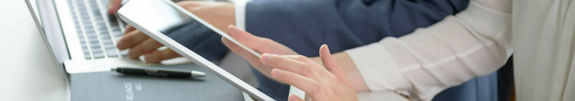Zwei Personen arbeiten an Laptop und Tablet in moderner Hotelumgebung, mit Notizbuch und Wasserglas auf dem Tisch.