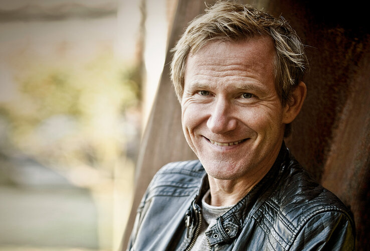 A person is smiling at the camera, wearing a black leather jacket, with blurred nature in the background.