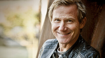 Eine Person lächelt in die Kamera, trägt eine schwarze Lederjacke, im Hintergrund unscharfe Natur.