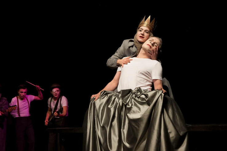 Theater scene with actor in crown embracing another; musician in the background. © bremer shakespeare company