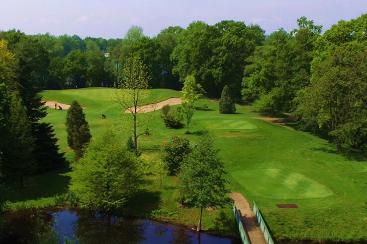 Golfplatz mit grünen Fairways, Sandbunkern, Bäumen und einem kleinen Teich im Vordergrund.