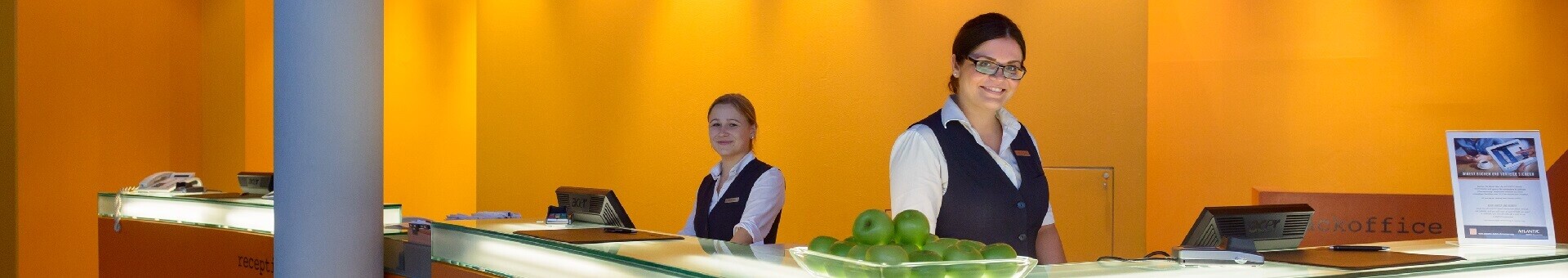Rezeption ATLANTIC Hotel Universum Bremen Hotelrezeption mit zwei lächelnden Mitarbeiterinnen, oranger Hintergrund, Glasplatte mit grünen Äpfeln.