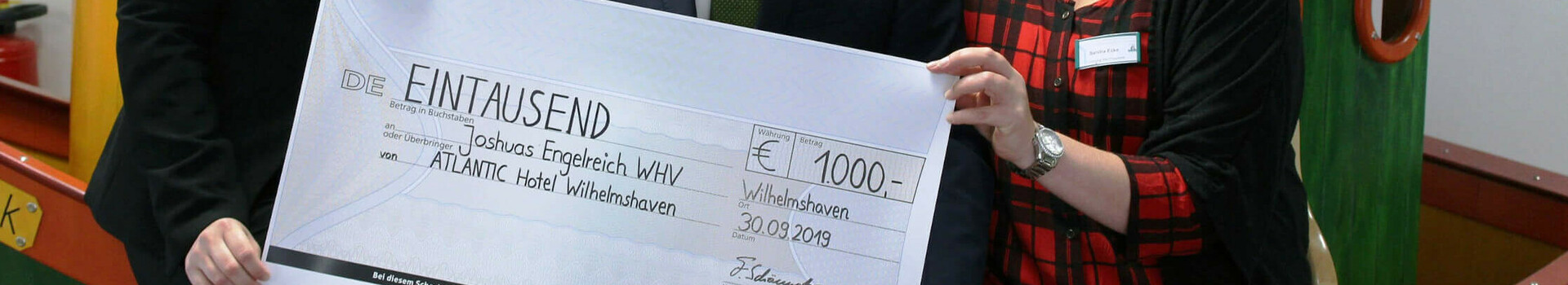 Spendenübergabe ATLANTIC Hotel Wilhelmshaven Drei Personen halten einen symbolischen Scheck über 1000 Euro im ATLANTIC Hotel Wilhelmshaven.