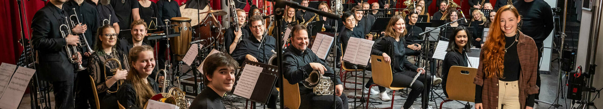 Christmas concert with the Ratekau Symphonic Wind Orchestra | ATLANTIC Grand Hotel Travemünde Orchestra in a rehearsal room with red curtains, musicians with instruments, a woman standing in front smiling.