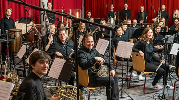 Orchester in einem Probenraum mit roten Vorhängen, Musiker mit Instrumenten, eine Frau steht lächelnd vorne.