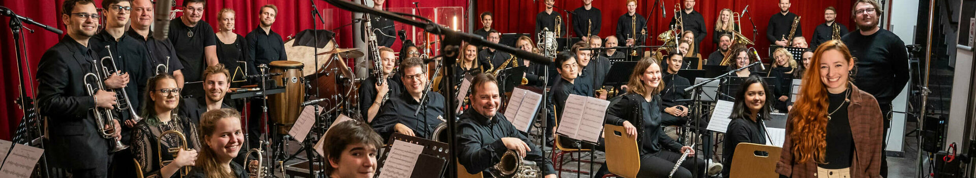 Orchester in einem Probenraum mit roten Vorhängen, Musiker mit Instrumenten, eine Frau steht lächelnd vorne.