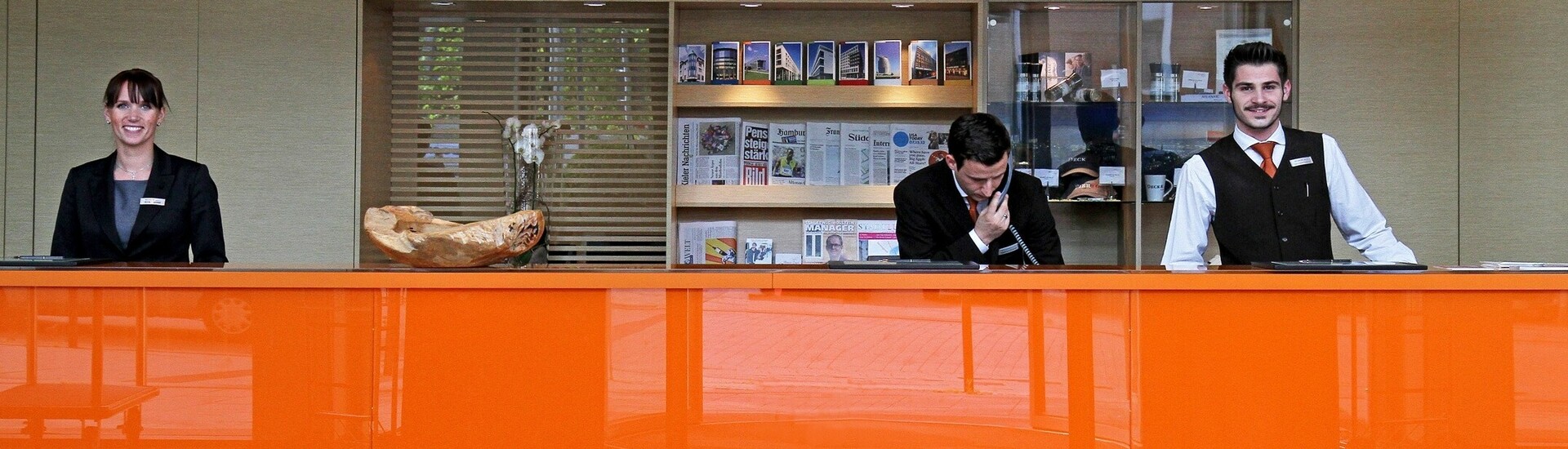 Hotelrezeption mit drei lächelnden Mitarbeitern hinter einem orangefarbenen Tresen.