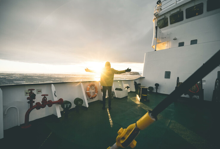 Person standing on a ship, arms outstretched as the sun sets over the sea.