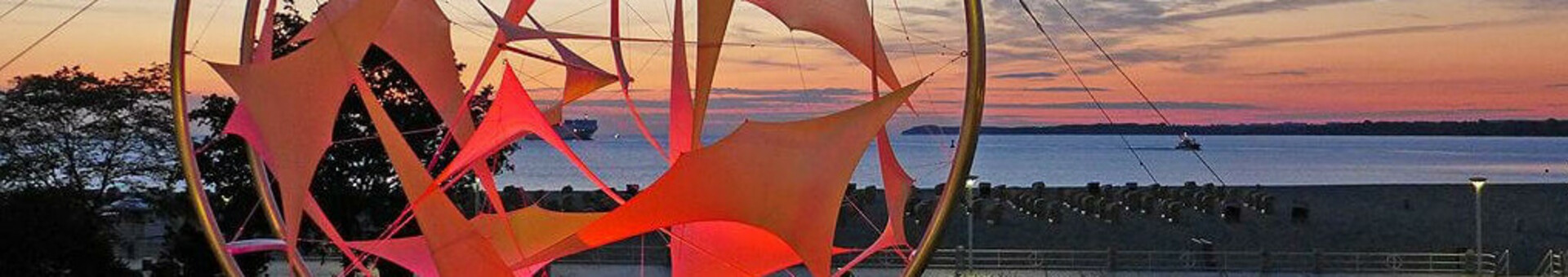 Leuchtende Skulptur am Strand von Travemünde bei Sonnenuntergang, mit Meer und farbigem Himmel im Hintergrund.