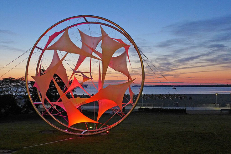 Leuchtende Skulptur am Strand von Travemünde bei Sonnenuntergang, mit Meer und farbigem Himmel im Hintergrund.