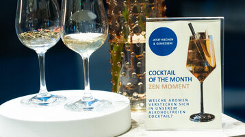 Aufnahme zweier Cocktailgläser, daneben ein Schild auf dem steht, "Cocktail of the Month: Zen Moment. Welche Aromen verstecken sich in unserem alkoholfreien Cocktail?"
