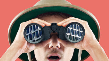 Person mit Safari-Hut schaut durch Fernglas, reflektiert Hotelgebäude, vor rotem Hintergrund.