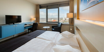 Bright hotel room with double bed, desk, TV and panoramic window with a view of Bremerhaven and the Weser.