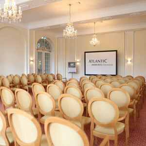 Tagungs- und Veranstaltungsraum "Bremen" Eleganter Konferenzraum mit Kronleuchtern, Reihen von Stühlen und einer Leinwand im ATLANTIC Grand Hotel Travemünde.